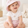 A smiling baby girl wearing a pink and white striped sun hat, a white short-sleeve top with floral embroidery, and matching pink striped shorts, sitting on a soft cream blanket with a stuffed bunny in the background.