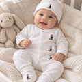 A smiling baby wearing a white footed onesie and matching beanie with small sailboat prints, sitting on a textured cream blanket next to a plush bunny toy.